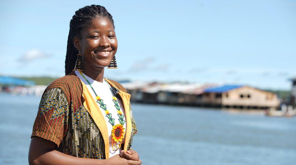 Imagen de mujer joven afro, lleva un collar con un girasol que resalta en su sonrisa, está frente a bahía en Buenaventura